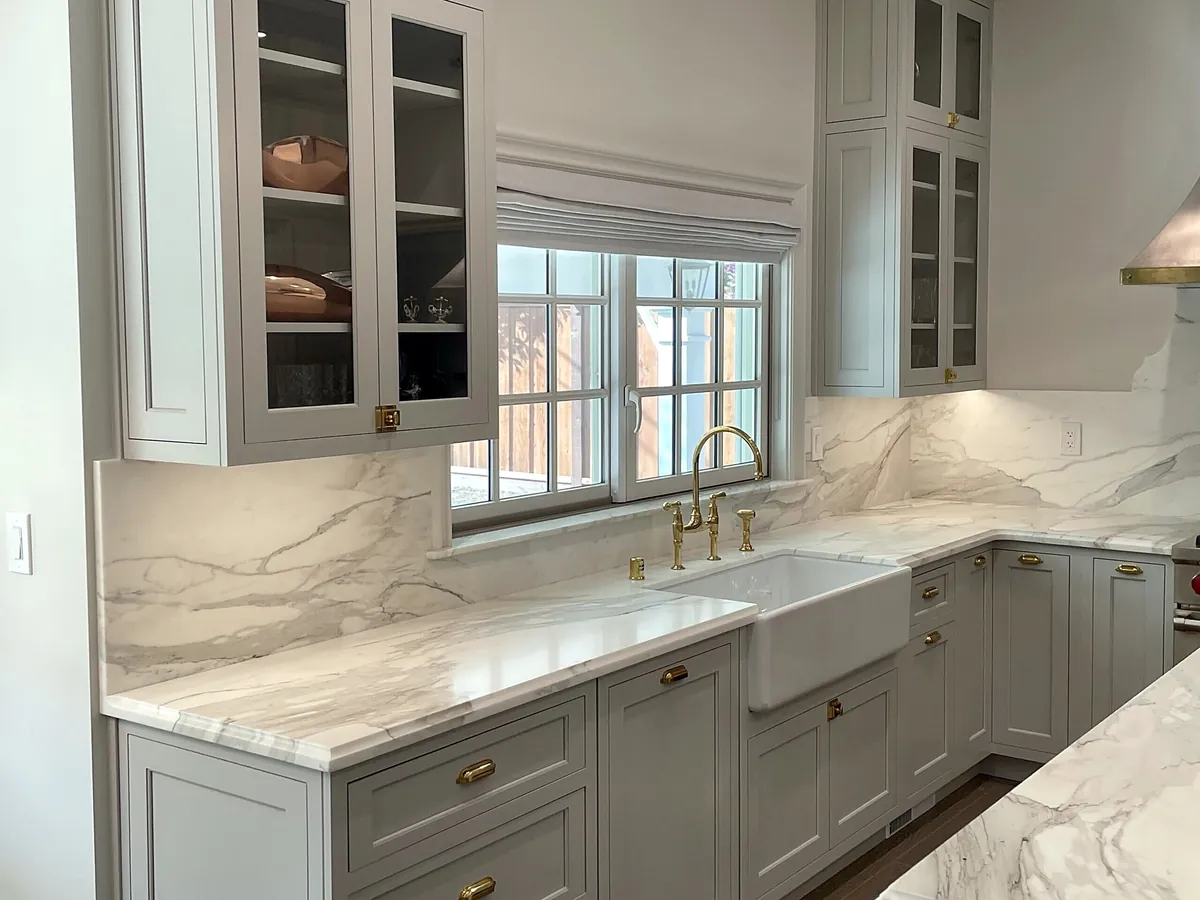 Farmhouse sink with brass faucet under a window, marble counters